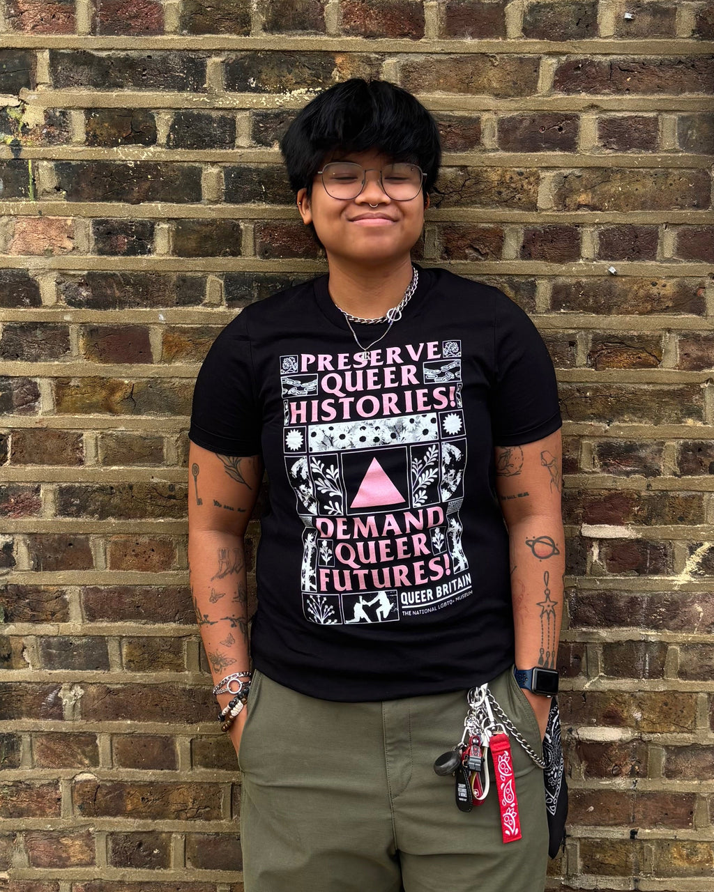 A person standing in front of a brick wall wearing a black graphic T-shirt with Queer History Month text and olive green pants.