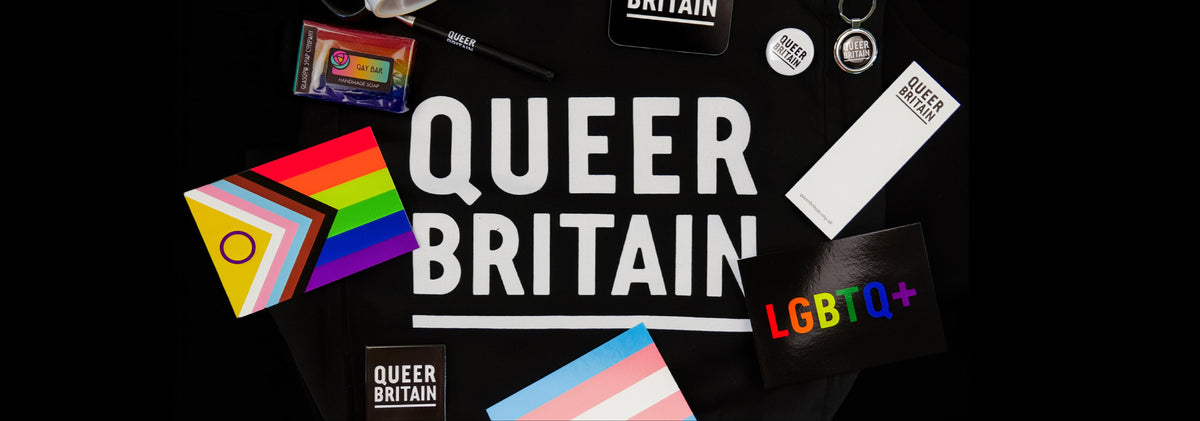A flat-lay arrangement of Queer Britain merchandise on a black background, including a white mug, stickers, a small cassette tape, a pen, a rectangular card, and a progress pride flag, with large QUEER BRITAIN text in the center and an LGBTQ+ symbol near the bottom right.